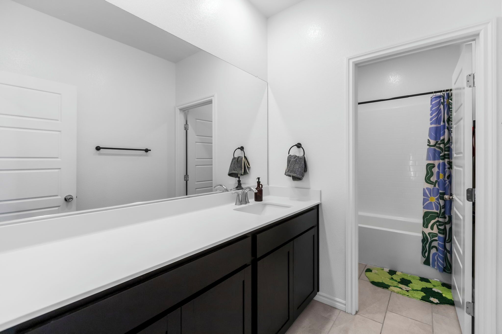 Un baño con lavamanos blanco, armarios negros, un espejo grande y una ducha con cortina estampada.