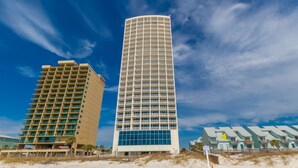 A tall, modern hotel with a curved facade, located on a sandy beach with a clear blue sky. - Island Tower Condos (Gulf Shores)
