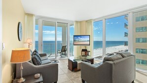 A modern living room with a large TV, comfortable seating, and a balcony with a view of the beach. - Island Tower Condos (Gulf Shores)