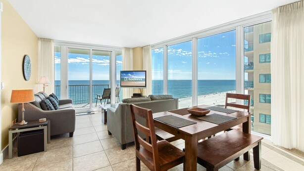 A spacious living area with a large window overlooking the ocean, a dining table with chairs, a sofa, and a television on the wall. - Island Tower Condos (Gulf Shores)
