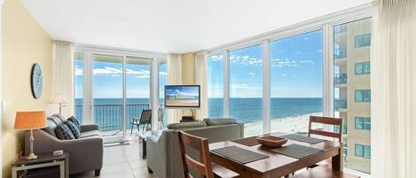 Ampla sala de estar com uma grande janela com vista para o oceano, uma mesa de jantar com cadeiras, um sofá e uma televisão na parede.
