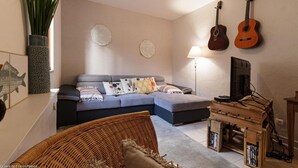 Ein Wohnzimmer mit einer Couch, einem Fernseher, zwei Gitarren, einer Lampe und einem Bücherregal.