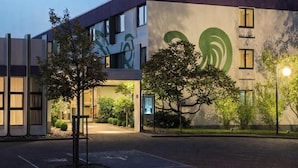 Een modern gebouw met een groot groen logo op de gevel, een parkeerplaats en bomen op de voorgrond.