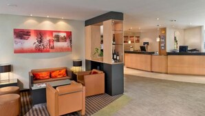 En modern hotellobby med en receptionsdisk, en sittgrupp med soffa och fåtöljer samt en stor, abstrakt målning på väggen.