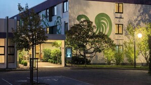 Un edificio moderno con un gran logo verde en la fachada, un área de estacionamiento y árboles en primer plano.