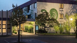 A modern building with a large green logo on the facade, a parking area, and trees in the foreground. - Sunday Saarbruecken Sued Deluxe Double Room (Saarbrücken)