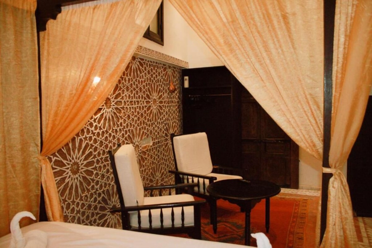 A room with two chairs and a small table, a patterned wall, and a bed with curtains.