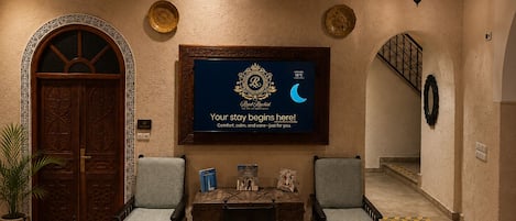 Um saguão de hotel com balcão de recepção, duas poltronas, um lustre e um letreiro de boas-vindas emoldurado.