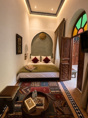 Un dormitorio con cama, un cabecero decorativo, un tapete estampado, una mesita con un control remoto y botellas de agua, y vistas a un espacio exterior con una puerta y un banco.