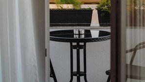 Vista desde una ventana a un balcón con una mesa redonda y sillas, plantas en macetas y un paisaje urbano de fondo.