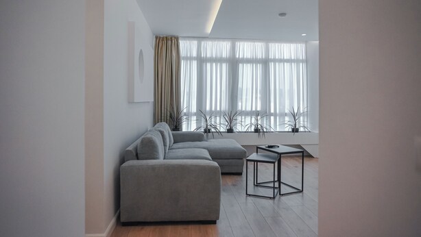 Una sala de estar moderna con un sofá seccional gris, una mesita negra y ventanas amplias con cortinas traslúcidas.
