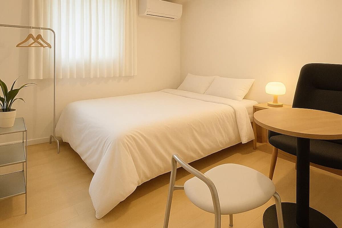 Une petite pièce bien éclairée, avec un lit simple, une chaise, une table, un porte-manteau mural et une plante en pot.