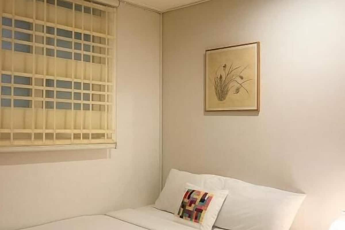 Une chambre à coucher avec un lit, une fenêtre avec des stores, une table de chevet et un tableau accroché au mur.