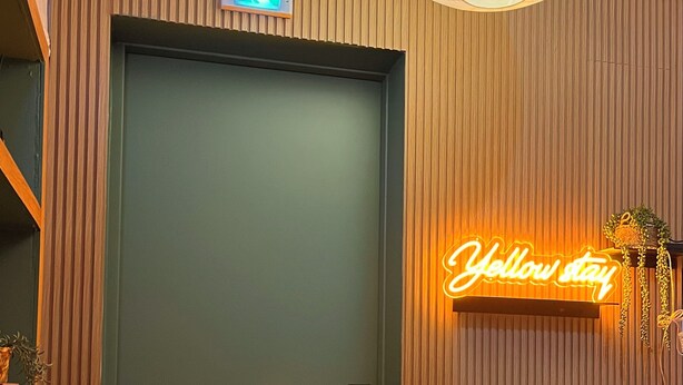 A hotel room with a wooden panel wall, a neon sign reading "Yellow stay", a door with a handle, and a small plant on the left side. - Yellow stay (Bucheon)