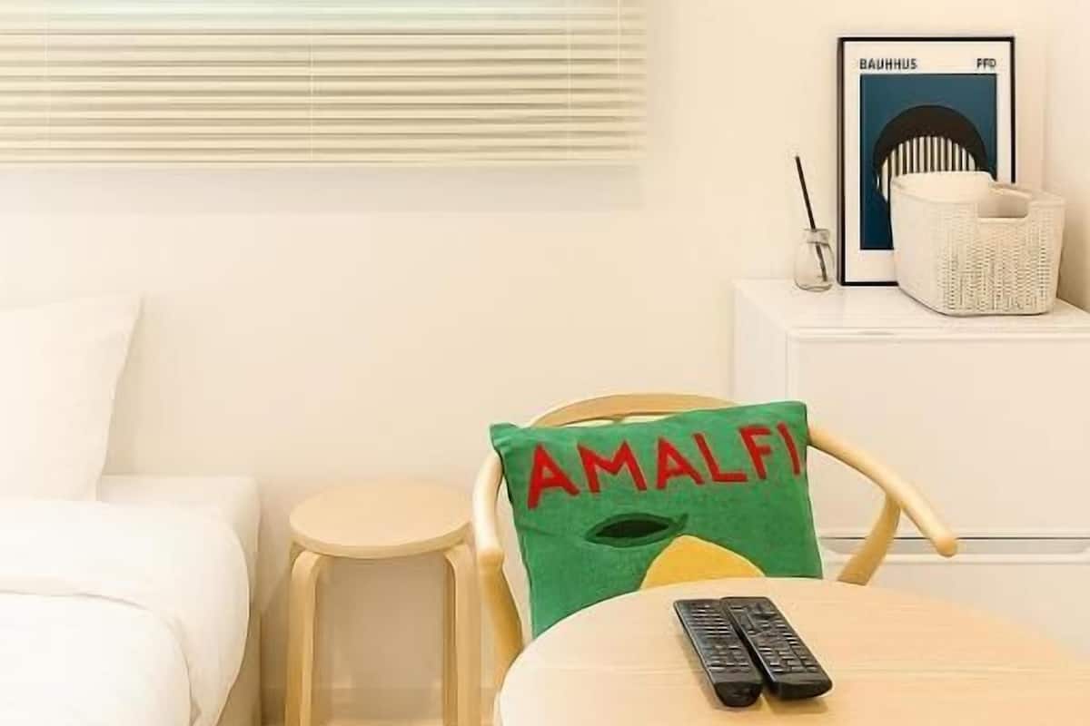 Une chambre d’hôtel avec un lit, une petite table ronde sur laquelle reposent deux télécommandes, un oreiller vert orné du mot « AMALFI », une table de chevet blanche avec une lampe, et un tableau encadré accroché au mur.