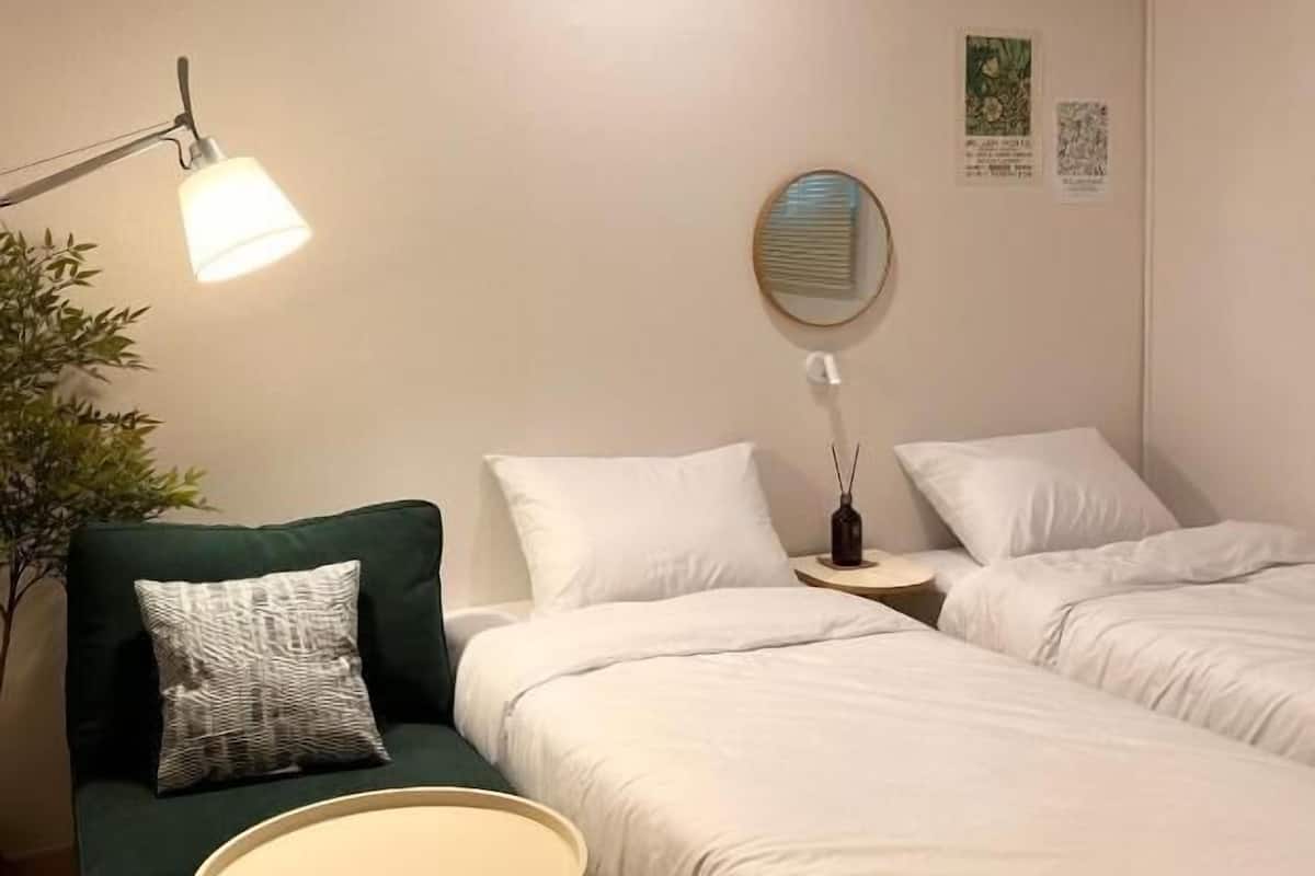Une chambre d’hôtel avec deux lits, un fauteuil vert, une petite table ronde et une lampe sur pied.