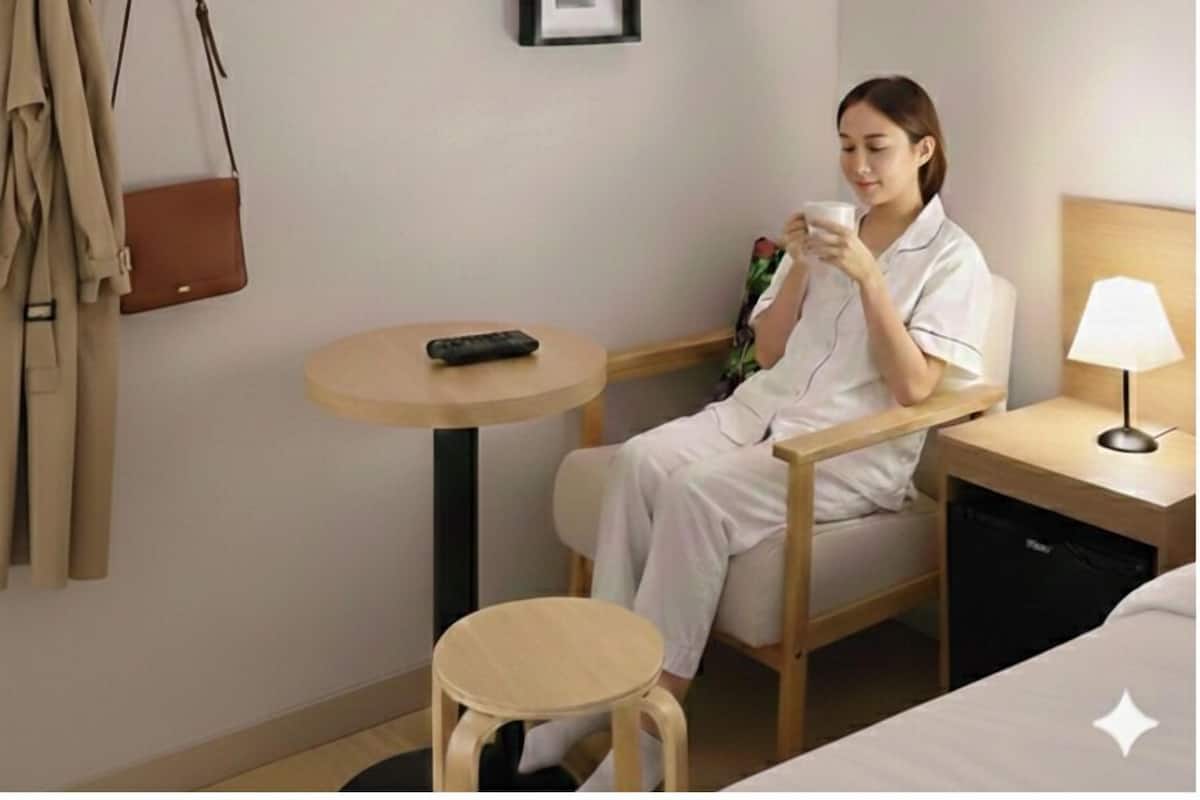 Une chambre d’hôtel avec un lit, une petite table ronde, une chaise, une table de chevet avec une lampe, un porte-manteau avec une veste, et deux tableaux encadrés accrochés au mur.