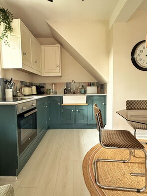 A kitchen with teal cabinets, a wooden dining table, and a wicker chair. - Stunning semi- rural cottage with a huge bed! (Wadhurst)