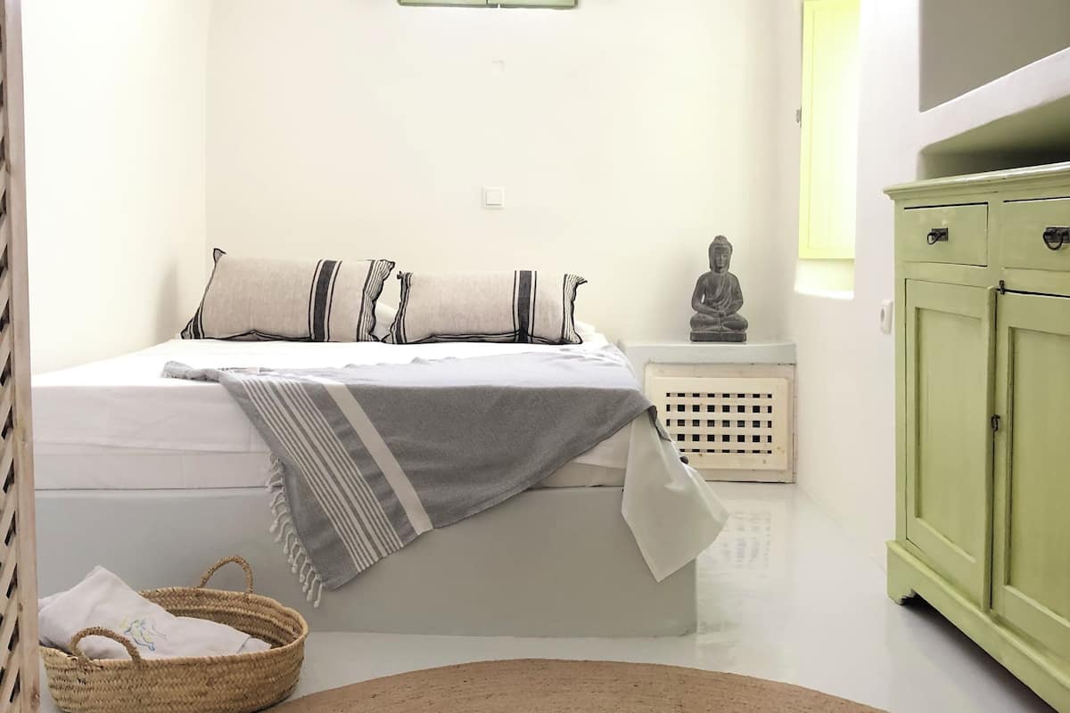 Een slaapkamer met een bed, nachtkastje, groen kastje, gevlochten mand en rond vloerkleed.