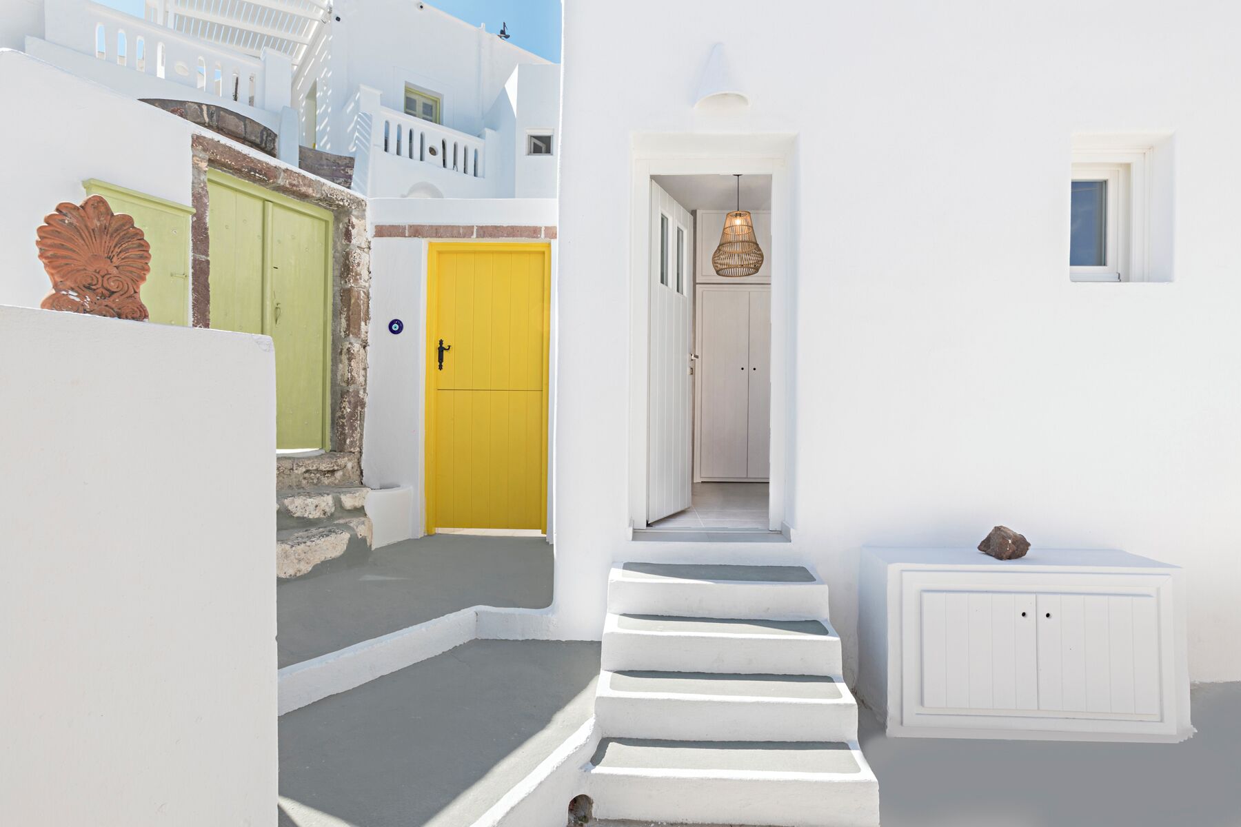 A narrow white-walled street with a yellow door and a staircase leading to a second-floor balcony.