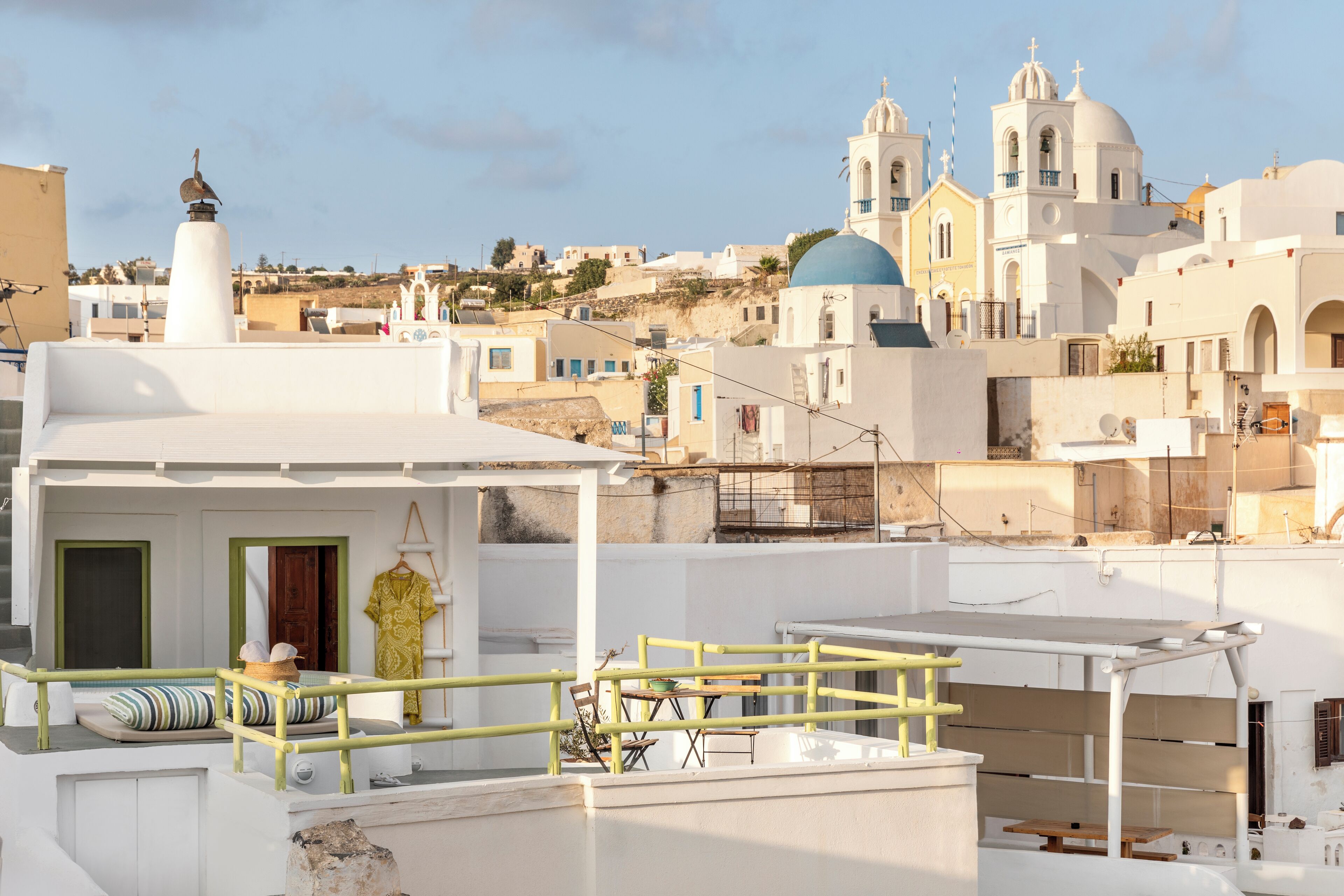 A rooftop terrace with a seating area, a small table, and a view of a coastal town with white buildings and blue domes.
