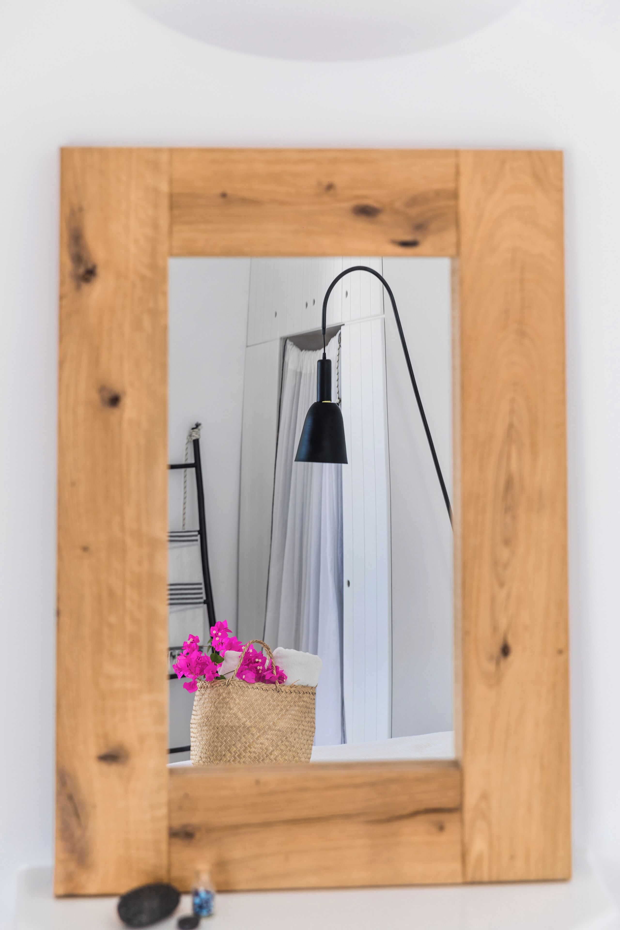 A framed mirror reflecting a room with a white wall, a black lamp, a wicker basket with pink flowers, and a white curtain.