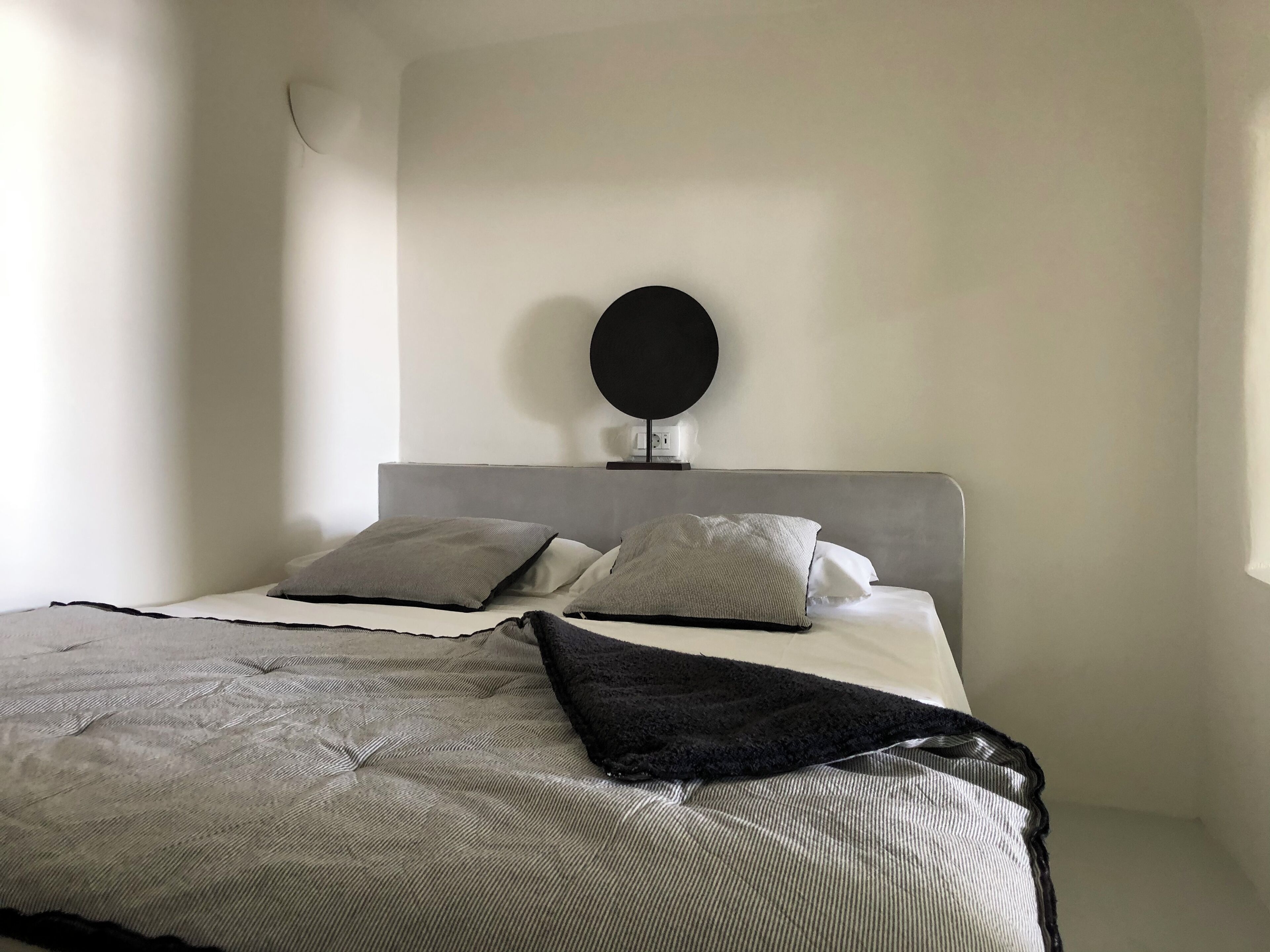 Un dormitorio minimalista con cabecera gris, dos almohadas grises, una manta de color negro y un espejo redondo en la pared.