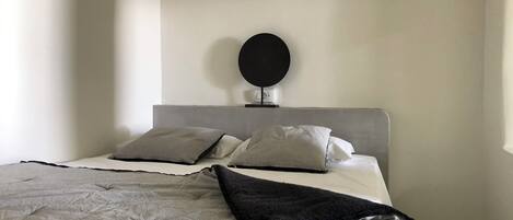 Un dormitorio minimalista con cabecera gris, dos almohadas grises, una manta de color negro y un espejo redondo en la pared.