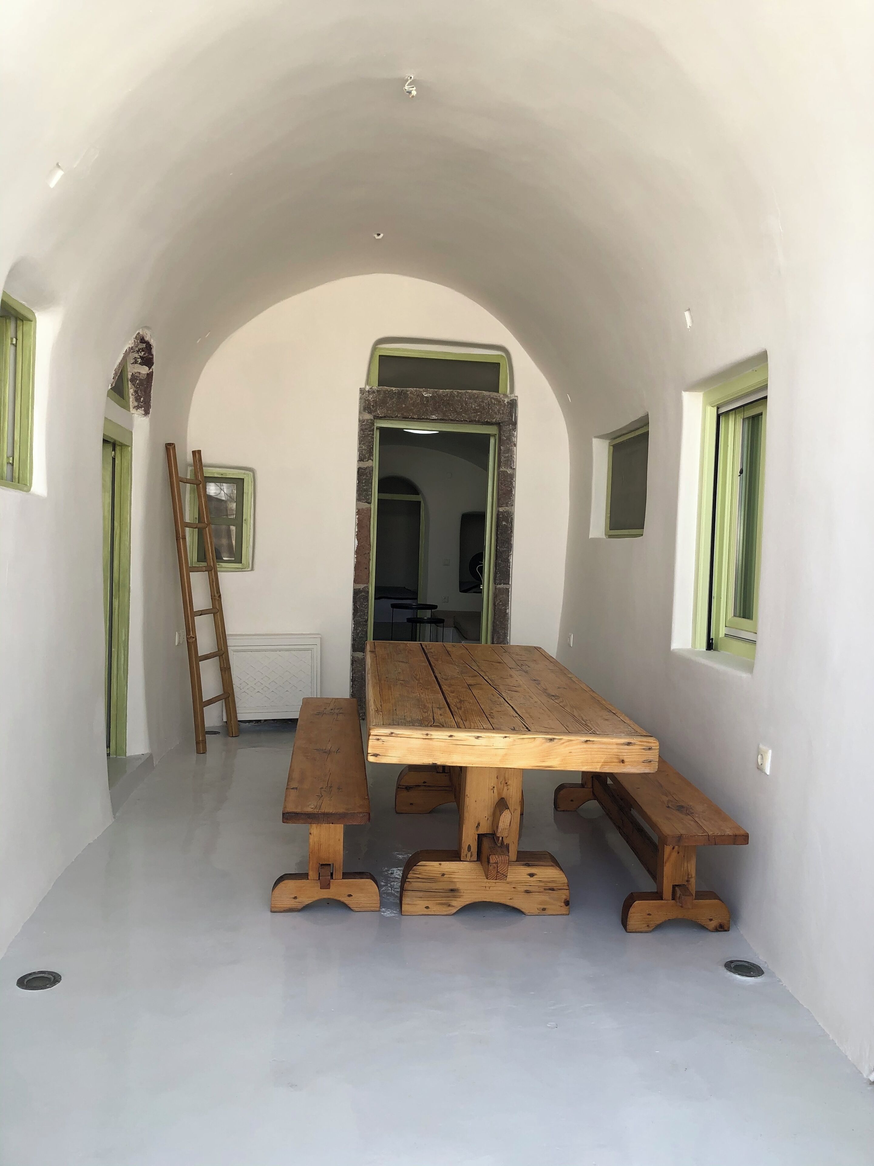 Un comedor con mesa y bancos de madera, techo abovedado y vista a otra habitación.