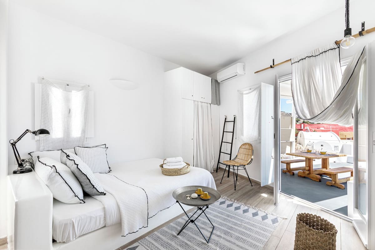 Een lichte, witte kamer met een bed, een klein tafeltje en een stoel. Er staat een ladder en een mand op de vloer.