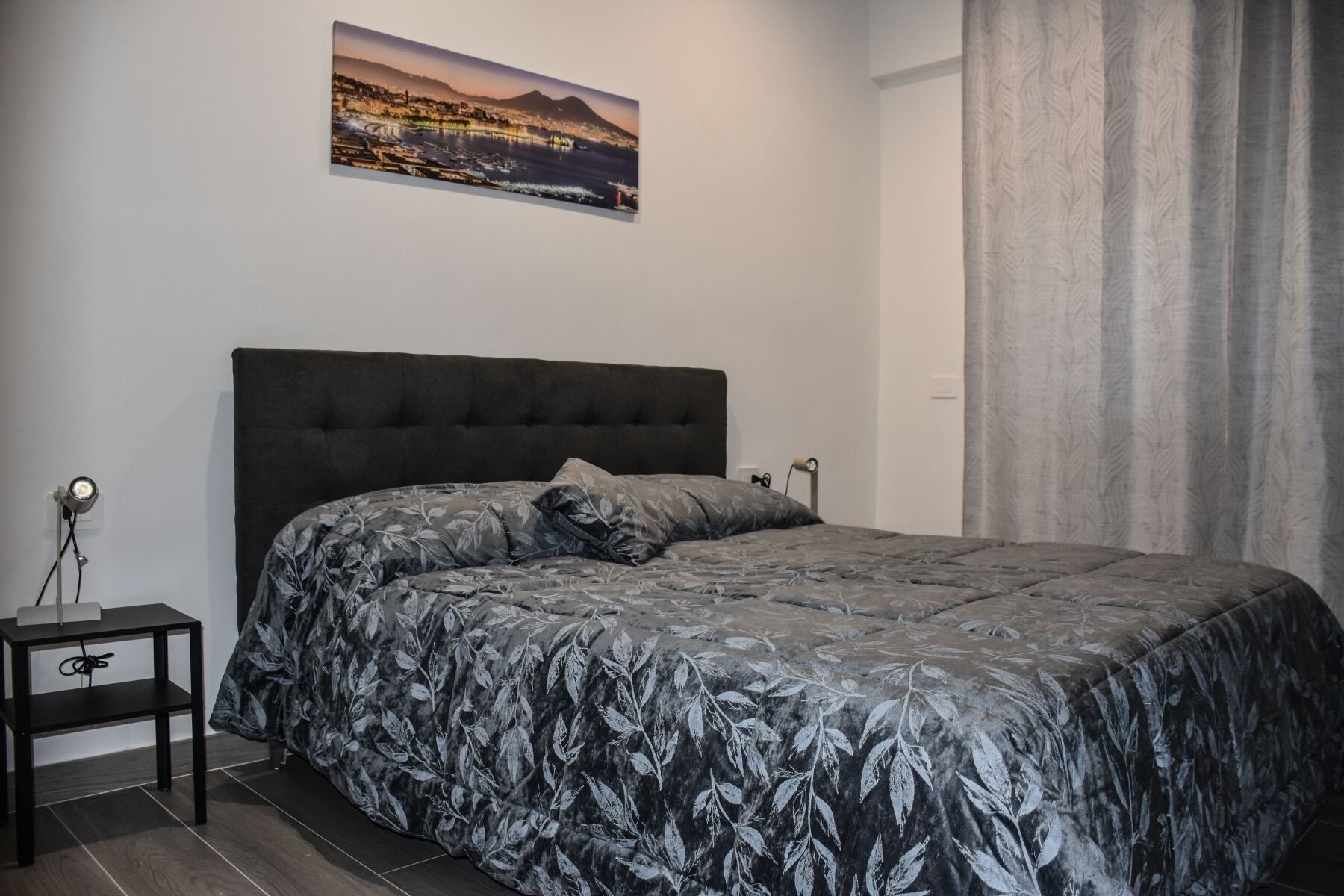 Un dormitorio con cama, mesita de noche, lámpara, un cuadro y una ventana con cortinas.