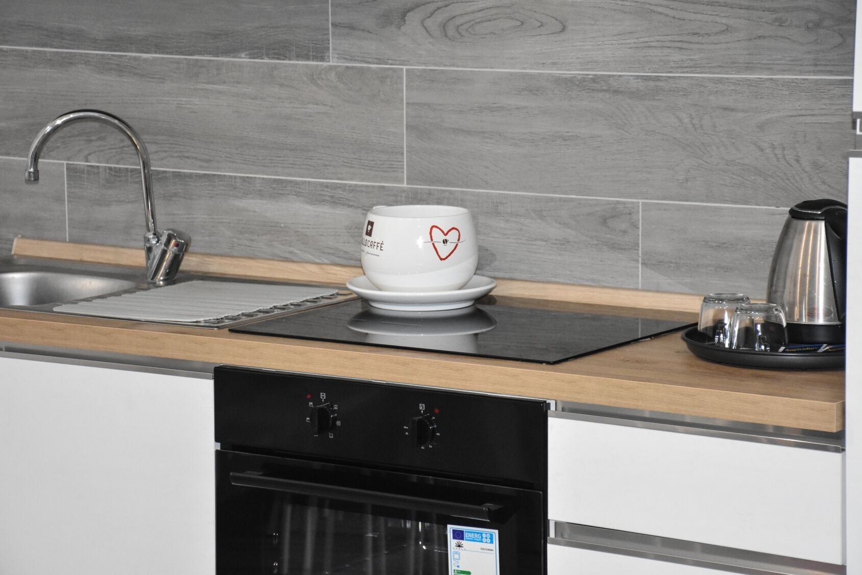 A modern kitchen with a stainless steel sink, a white coffee cup with a heart design, a kettle, and a black tray with glasses.