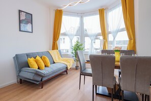 Una sala de estar moderna con un sofá gris, cojines amarillos, una mesa de comedor con sillas y un ventanal con cortinas amarillas.