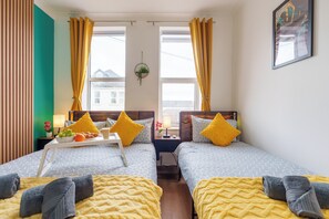 Habitación doble con dos camas, cada una con una manta y almohada amarillas, una mesita con fruta y una lámpara, y una ventana con cortinas.
