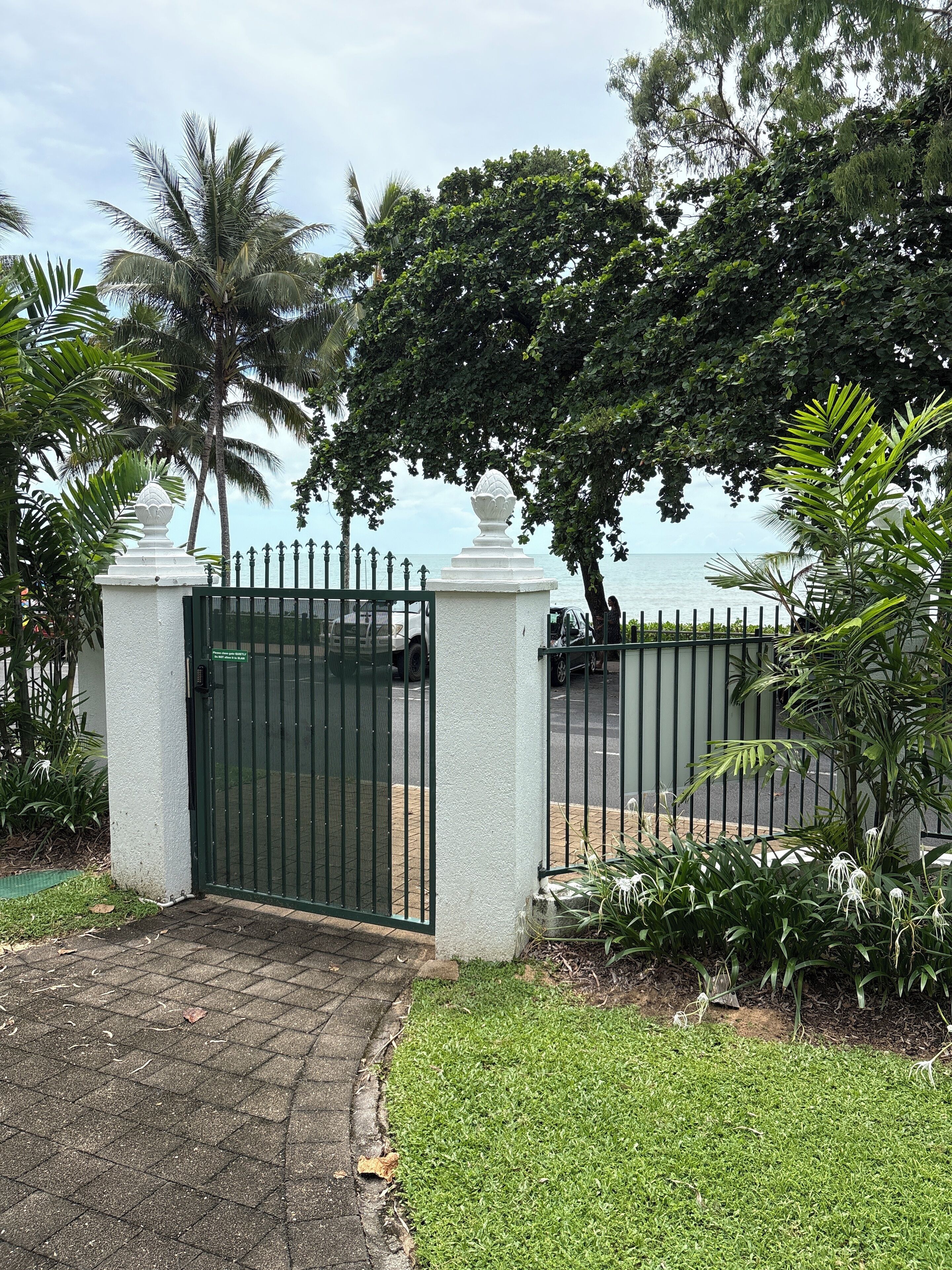 A gated entrance with a paved path leading to a lush garden area, palm trees, and a view of the sea.