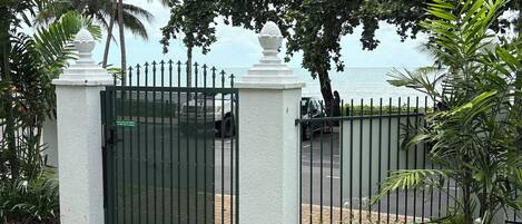 A gated entrance with a paved path leading to a lush garden area, palm trees, and a view of the sea.