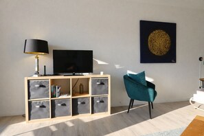 Ein modernes Wohnzimmer mit einem Flachbildfernseher auf einem Holztv-Schrank, einem türkisfarbenen Sessel und einem an der Wand hängenden Kunstwerk.