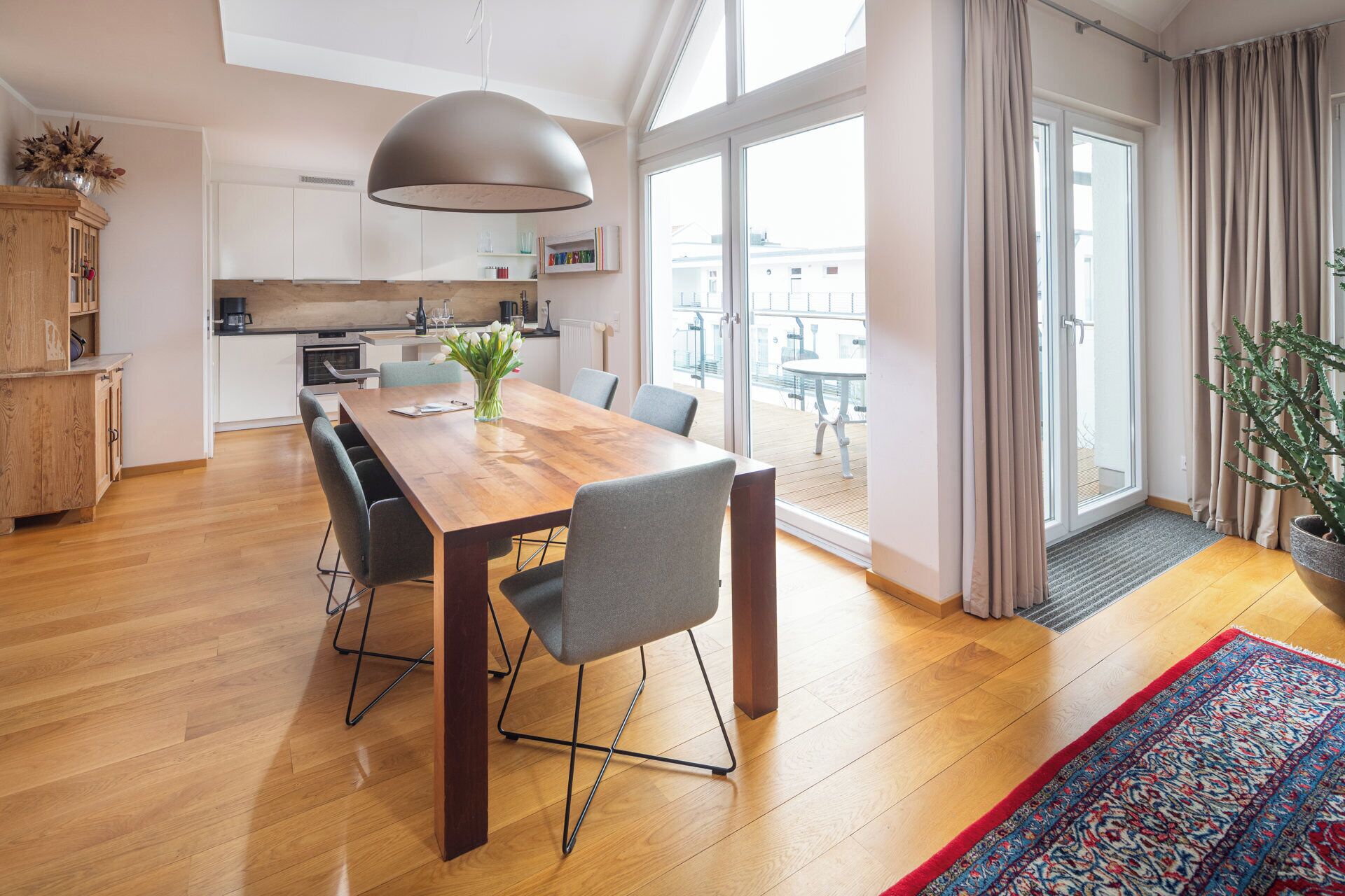 A modern apartment with a wooden dining table, grey chairs, a large pendant light, a kitchen with white cabinets, and a balcony with a view.