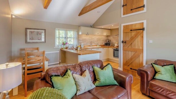 A spacious living area with a leather sofa, a dining table, and a kitchen in the background. - The Gate Lodge - One Bed Cottage - Pet Free - Nr Finchingfield & Saffron Walden (Little Bardfield)