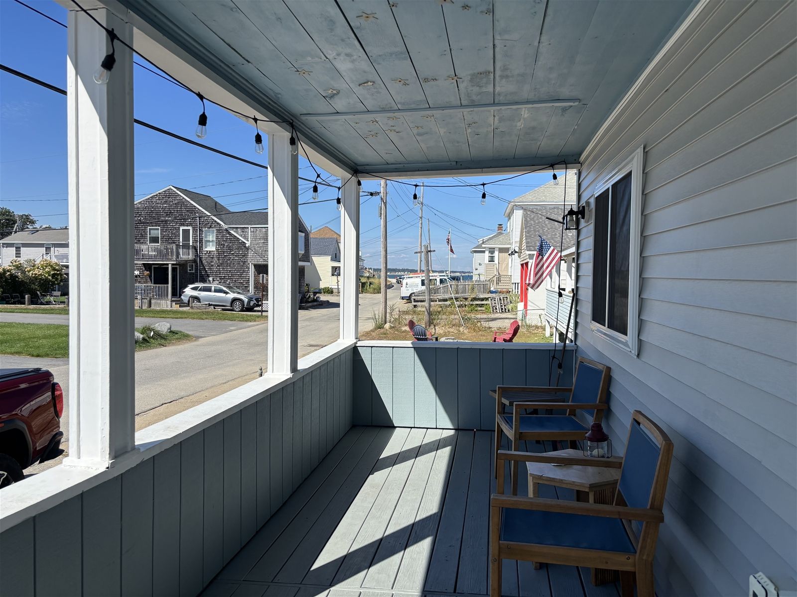 A covered porch with wooden chairs and a table, overlooking a street with houses and parked cars.