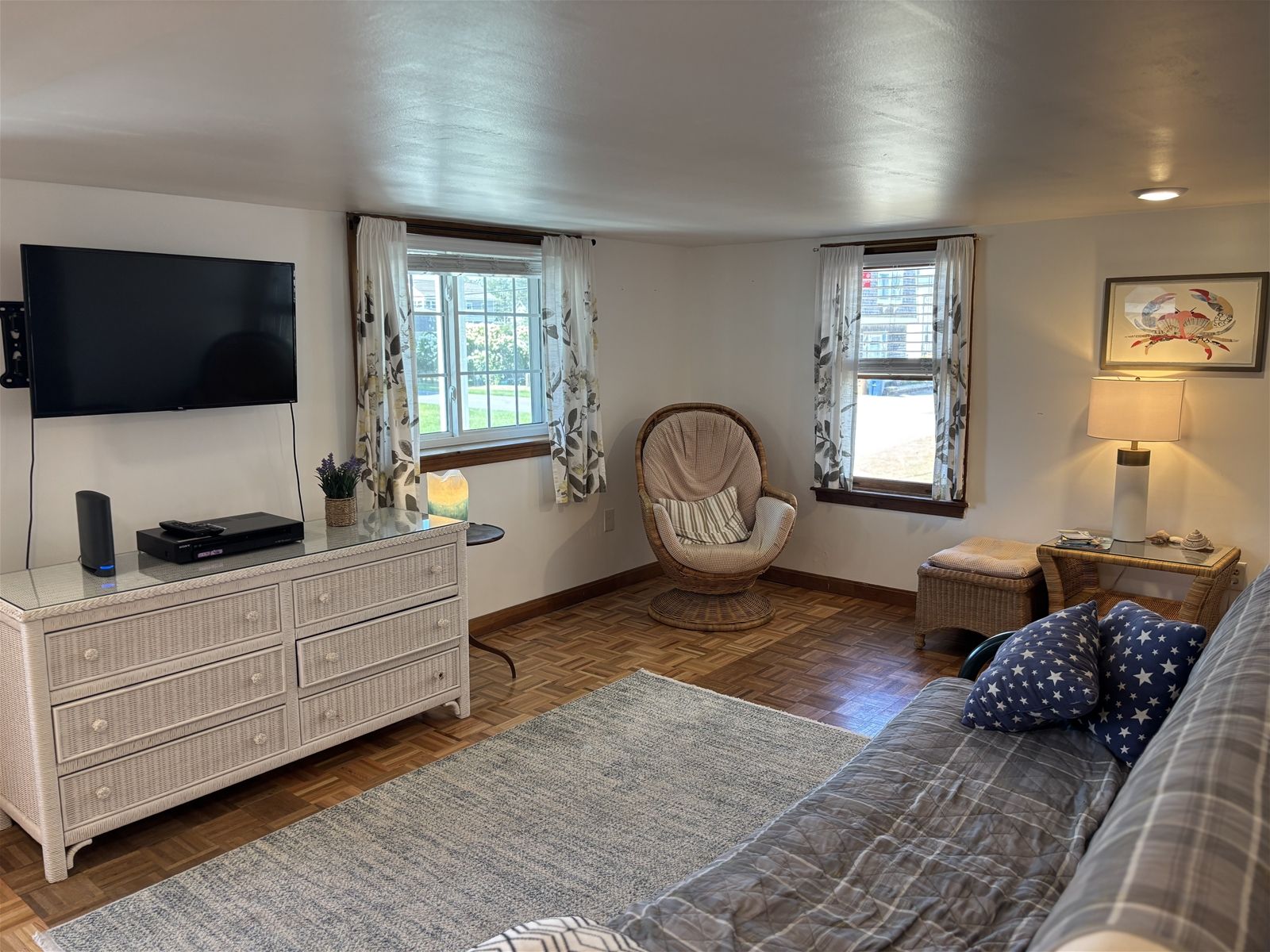 A cozy room with a flat-screen TV, a wicker dresser, a cushioned chair, a sofa with star-patterned pillows, and a small table with a lamp.