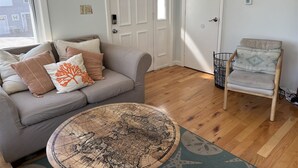 A living room with a grey sofa, a round coffee table with a map, a wooden chair, and a white door. - Experience coastal charm at Sand Dollar, a spacious 4-bedroom retreat in Ferry Beach, Saco, ME. (Saco)