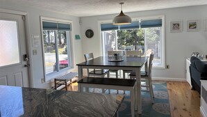 A dining area with a table and chairs, a pendant light, and a view of the outdoors through large windows. - Experience coastal charm at Sand Dollar, a spacious 4-bedroom retreat in Ferry Beach, Saco, ME. (Saco)