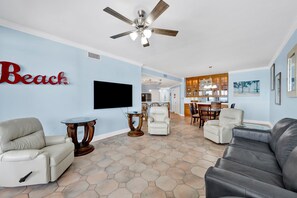 A living room with a TV, two leather sofas, a ceiling fan, and a dining area with chairs and a table. - Dog Friendly Beachfront Paradise East Corner Unit (Orange Beach)