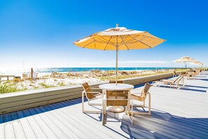 A wooden deck with outdoor seating, including chairs and tables, overlooking a beach with sand dunes and the ocean. - Dog Friendly Beachfront Paradise East Corner Unit (Orange Beach)