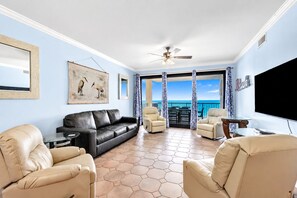 A living room with a black leather sofa, beige armchairs, a glass coffee table, a ceiling fan, and a large TV. - Dog Friendly Beachfront Paradise East Corner Unit (Orange Beach)