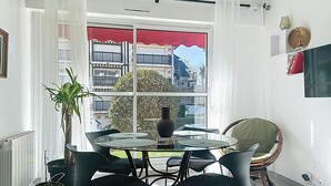 Un coin repas avec une table en verre et des chaises noires, une plante en pot et une vue sur les bâtiments à travers une grande fenêtre.