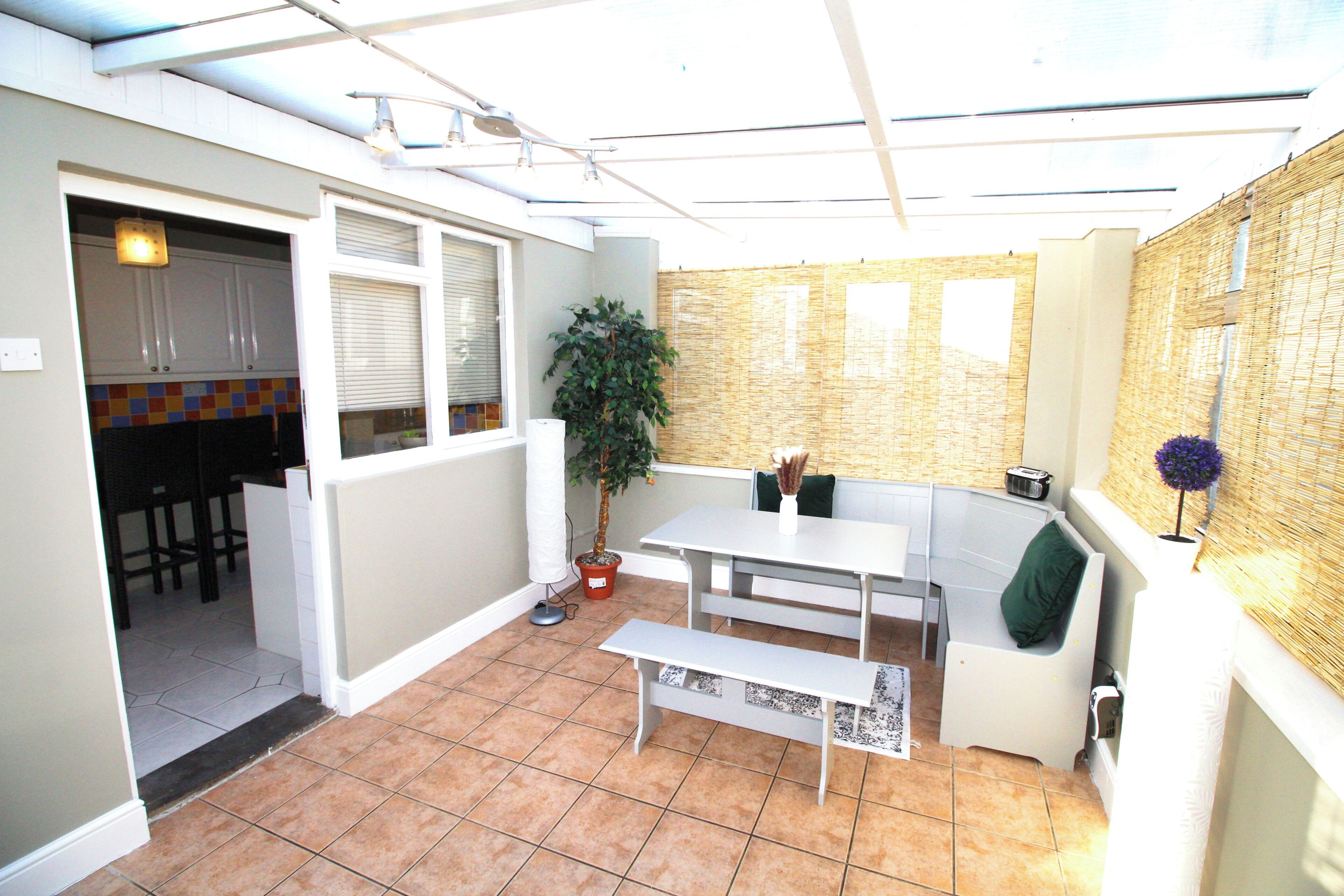 A compact kitchen with white cabinets, a dining table, and a bench. There is a potted plant and a small table with a decorative item.