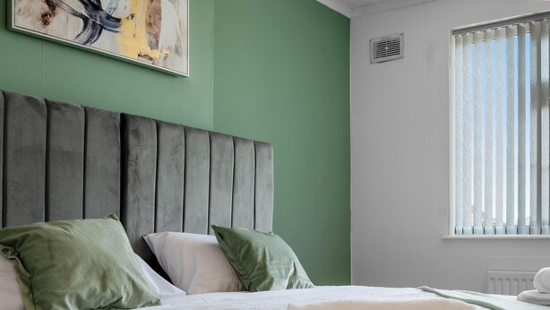 Een hotelkamer met een bed, kussens, een hoofdbord, een raam met zonwering en een schilderij aan de muur.
