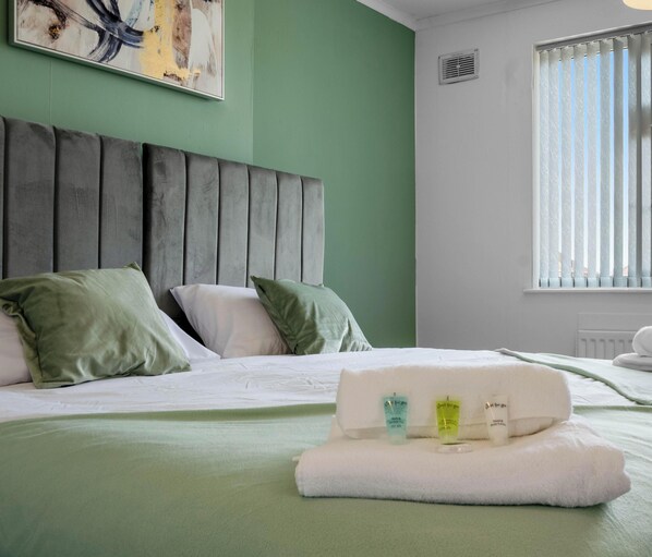 A hotel room with a bed, pillows, a headboard, a window with blinds, and a painting on the wall.